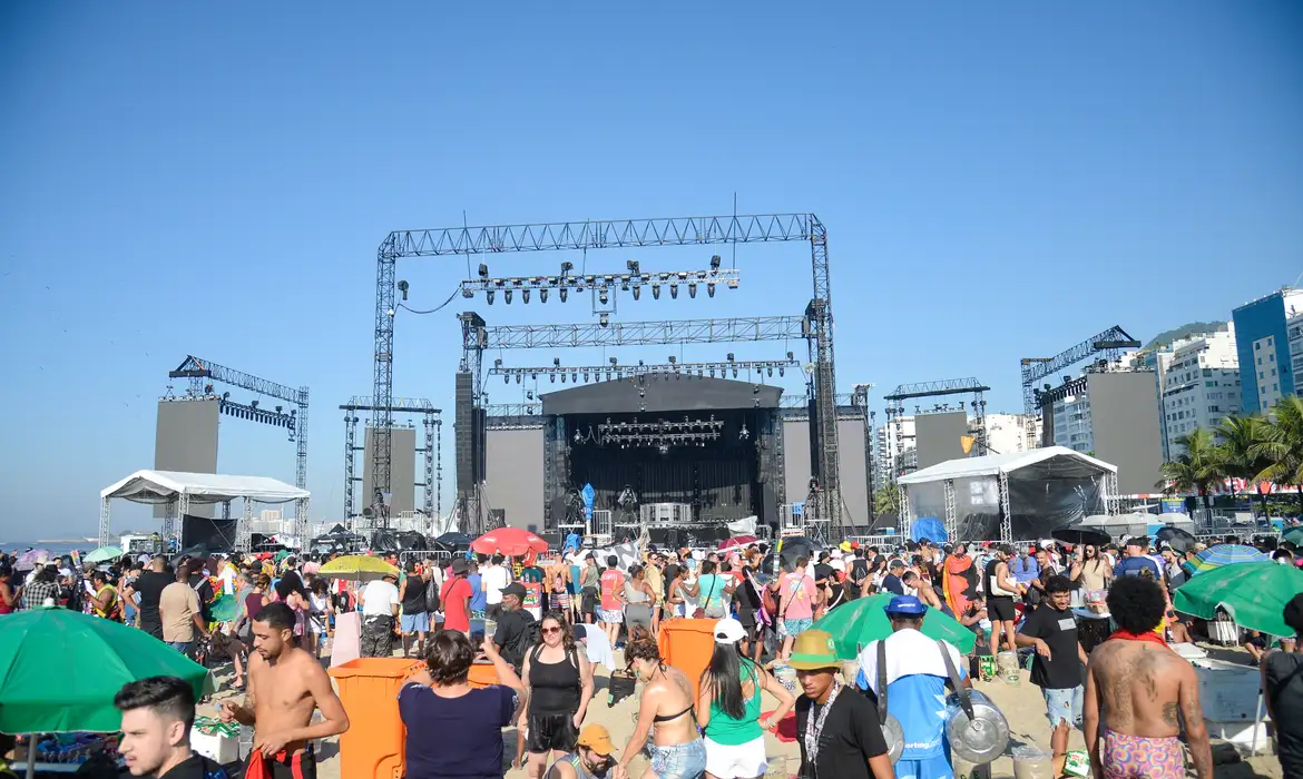 Ainda era pouco antes das 9h deste sábado (4), quando dezenas de pessoas correram afobadas para se posicionar na grade em frente ao palco do show da cantora Madonna, que começa às 21h45. O espaço era mantido isolado, por tapumes, desde o início dos trabalhos de montagem da megaestrutura, erguida nas areias da praia de Copacabana, na zona sul da cidade do Rio de Janeiro, para marcar o encerramento da turnê mundial da artista norte-americana. A turista argentina Gloria González mantinha seu lugar próximo aos tapumes desde a noite de sexta-feira (3). Ela chegou ao Rio no meio da semana, especialmente para acompanhar o show da cantora, de quem é fã mas a quem nunca assistiu ao vivo. Mesmo após a retirada dos tapumes, segurança ainda mantinham um cordão de isolamento. Quando eles permitiram que os fãs se aproximam da grade, os mais atentos conseguiram correr antes e se posicionar melhor. Gloria estava distraída e acabou chegando depois. Por sorte, a argentina fez amizade com uma brasileira que conseguiu reservar um lugar em que as duas conseguiram ficar encostadas na grade. Rio de Janeiro (RJ), 04/052024 – A turista argentina, Glória González em frente ao palco enquanto aguarda para show da Madonna, que está programado para a noite de hoje (04). Foto: Tomaz Silva/Agência Brasil A turista argentina Glória González em frente ao palco enquanto aguarda para show da Madonna. Foto: Tomaz Silva/Agência Brasil “Vamos ficar aqui até a hora do espetáculo, porque provavelmente é a minha última oportunidade de vê-la. Depois não sei onde ela vai se apresentar e nem se ela vai se apresentar de novo”, disse a turista, que se protegia do sol já forte com um chapéu preto, onde se lia “Madonna no Rio: eu fui”. A fã mineira Adriana Cruzeiro veio para o Rio de Janeiro com o marido em um ônibus fretado. Chegou por volta das 5h30 da manhã e já buscou um lugar junto às grades, que nem são tão perto do palco, uma vez que há uma área VIP, para a qual o acesso é restrito a convidados dos organizadores, no meio do caminho. “Estou disposta a ficar aqui até o final, porque outra oportunidade dessa, não vou ter nunca mais na minha vida. Vim com meu marido e três funcionários dele. Vamos ficar revezando aqui, para guardar o lugar até o show, mas depois de um certo horário, acho que vai ser impossível sair daqui”. Rio de Janeiro (RJ), 04/052024 – O turista George Oliveira em frente ao palco enquanto aguarda para show da Madonna, que está programado para a noite de hoje (04). Foto: Tomaz Silva/Agência Brasil O turista George Oliveira em frente ao palco enquanto aguarda para show da Madonna, que está programado para a noite de hoje (04). Foto: Tomaz Silva/Agência Brasil O paulista George Oliveira não faz questão de estar colado na grade, mas já tinha reservado um lugar, junto com o namorado e amigos, próximo dali. Sua estratégia para ver Madonna de perto? Um binóculo que pegou emprestado com o irmão. “Quero ver ela cantando, mexendo a boca. Se eu conseguir ver isso com o binóculo, já ganhei meu dia”, conta o esperançoso fã. Outra estratégia usada para visualizar melhor o palco é subir em um banquinho. O ambulante Adálisson Pereira anunciava a venda do banco, apelidado de “camarote da Madonna”, por R$ 40. “Já vendi mais de 100 desde ontem. Por enquanto o preço está R$ 40, mas vai subir mais perto do show”. Show A prefeitura do Rio de Janeiro estima um público de 1,5 milhão na noite deste sábado, em Copacabana. O espetáculo da cantora, que começa às 21h 45, terá duas horas de duração. O evento será aberto com DJ Barata, a partir das 18h45, e Diplo, às 20h, em um palco secundário, localizado próximo ao principal. Logo após o show de Madonna, haverá apresentação do DJ Pedro Sampaio. Mais de 3 mil policiais devem garantir a segurança do evento, que contará com pontos de revista e câmeras de reconhecimento facial. O bairro será fechado a veículos particulares a partir das 18h e a ônibus e táxis, a partir das 19h30. As melhores opções para chegar ao show são o metrô e os ônibus.